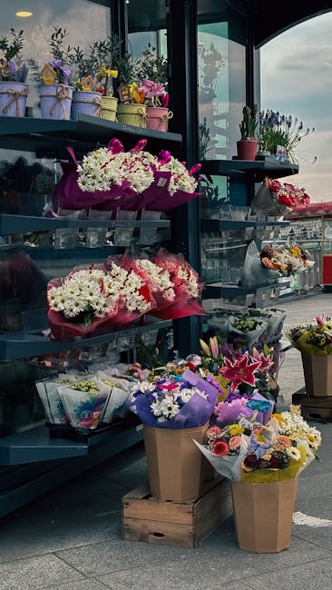 A display of various floral arrangements and bouquets on shelves and the ground outside a flower shop. The flowers include white daisies, pink lilies, and purple orchids, wrapped in colorful paper and