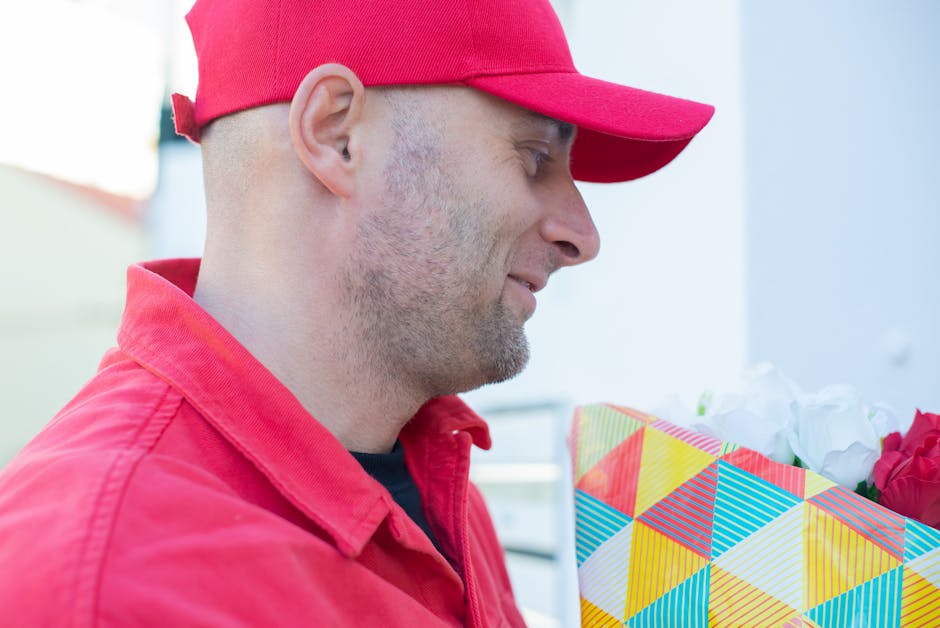 A delivery person wearing a red cap and red jacket is holding a colorful floral gift box with geometric patterns in yellow, red, blue, and white. The box contains fresh white and pink roses, with some