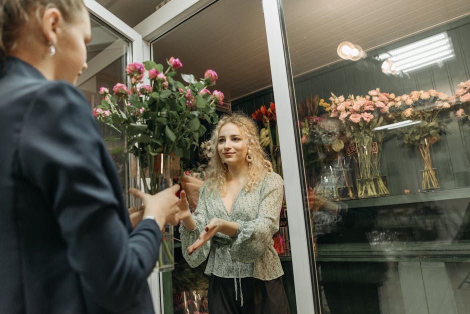 A young woman with curly, blonde hair and fair skin, dressed in a light green, floral-patterned blouse with long sleeves, is standing inside a flower shop near the glass window display. She is receivi