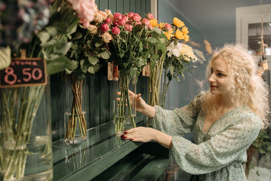 A woman with curly blonde hair and light skin selects a bouquet of flowers at a florist shop, [COMPANY_NAME], on Bellegrove Road. She is wearing a light, patterned blouse and has red painted nails. Th