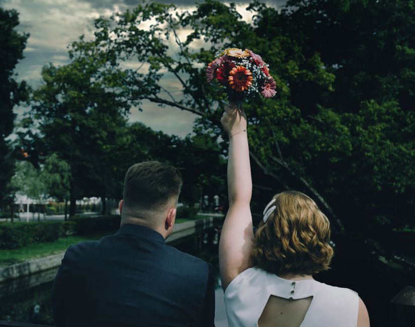 A woman with shoulder-length curly red hair, dressed in a white dress, holds a vibrant bouquet of flowers high above her head during a wedding event, viewed from behind. The bouquet features large ora