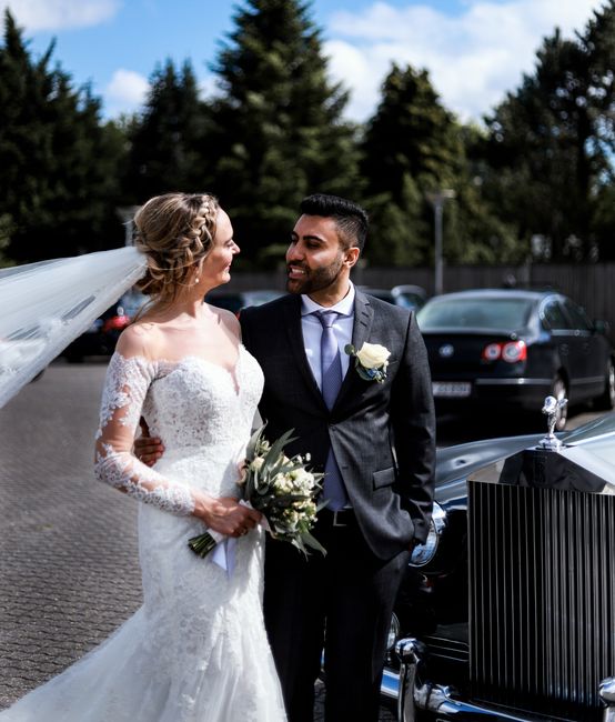 A bride and groom stand outdoors in a parking area during their wedding, with the bride wearing a white lace wedding gown with long sleeves and an off-the-shoulder neckline, holding a bouquet of white