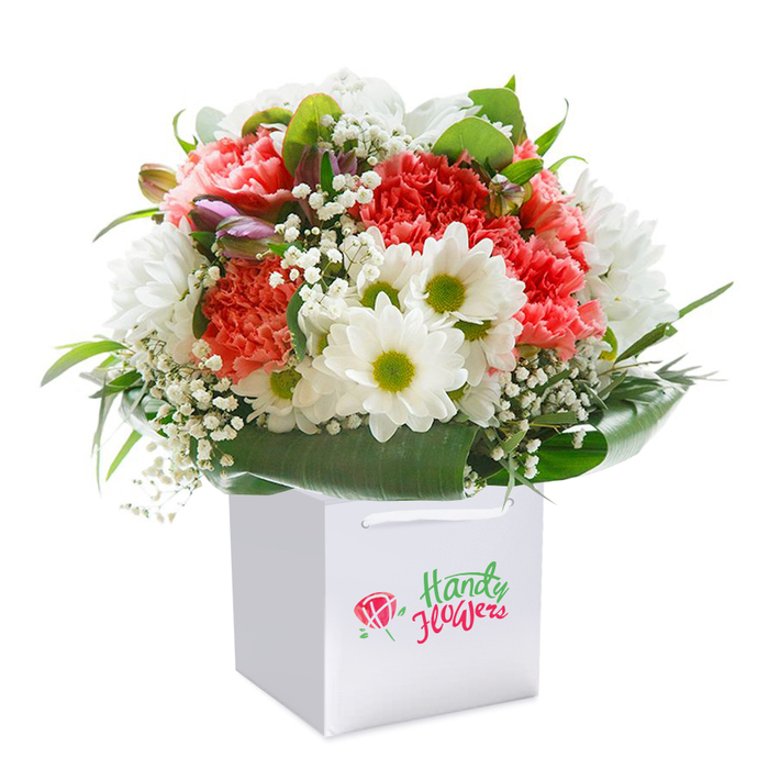 A lush, uplifting bouquet sits proudly within a crisp white gift bag, its vibrant blend of flowers reminiscent of the colourful borders along Welling's Bellegrove Road. The arrangement showcases soft coral, lively peach, and bold red carnations, their characteristic ruffled petals bursting with joyful energy. Bright white daisy-like blooms provide a spirited, fresh contrast, each centered by a sunny green heart that seems to capture a bit of spring's own delight. Ethereal clusters of baby's breath lace through the display, evoking gentle clouds and a sense of purity. Tucked throughout the arrangement, you'll spot delicate purple-tinged buds-possibly alstroemeria-suggesting the excitement of blooms yet to fully open. Framing it all is a lush collar of deep, polished green foliage that lends fullness, structure, and sophistication. The bouquet's carefully rounded composition, framed by sturdy white ribbon handles atop the bag, ensures a graceful silhouette and ease of gifting-ideal for celebrations, get-well wishes, or thoughtful surprises delivered across Welling and surrounding areas. Photographed against a seamless white backdrop, the flowers radiate freshness and craftsmanship, truly capturing the heartfelt quality delivered by passionate local florists.